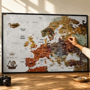 May include: A framed wall map of Europe featuring a vintage-style design with muted earth tones. The map includes text labels for countries and bodies of water, with the title Travel Story. It is decorated with numerous colorful push pins.