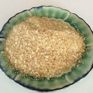 Può includere: Primo piano di una piccola ciotola di ceramica verde e blu piena di un cumulo di alghe secche di colore marrone chiaro granulare.