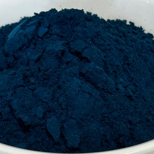 May include: Close-up of a pile of finely ground, deep blue powder. The powder has a textured appearance, with visible granules and a slightly uneven surface. The color is a rich, dark shade of blue, suggesting a pigment or dye.