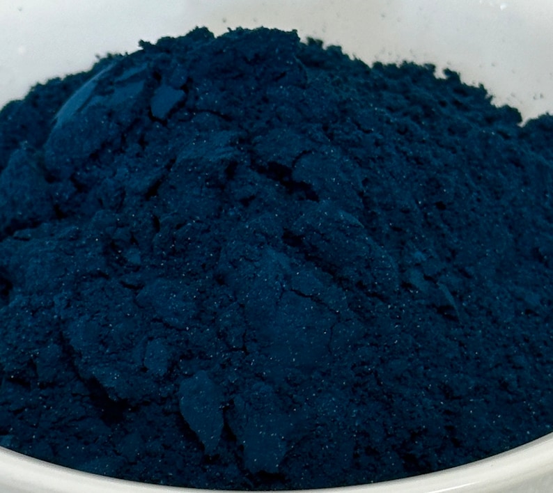 May include: Close-up of a pile of finely ground, deep blue powder. The powder has a textured appearance, with visible granules and a slightly uneven surface. The color is a rich, dark shade of blue, suggesting a pigment or dye.