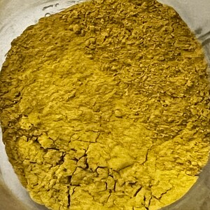 May include: Close-up of a jar filled with a finely ground, yellowish-brown powder. The powder has a textured, cracked appearance, suggesting it is dry. The image is taken from a top-down perspective, showing the full circular shape of the powder within the jar.