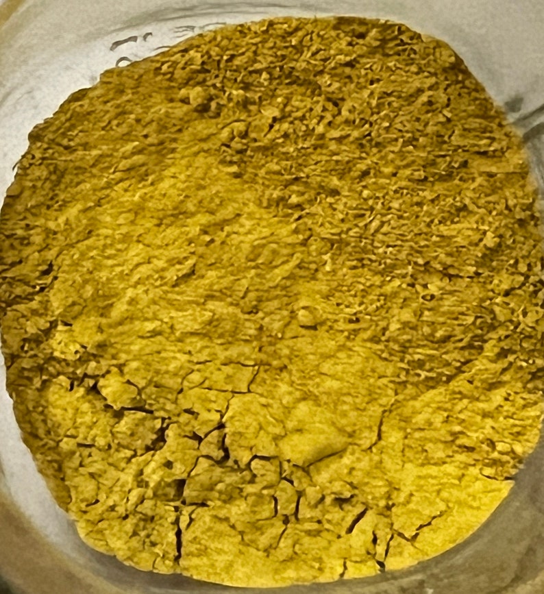 May include: Close-up of a jar filled with a finely ground, yellowish-brown powder. The powder has a textured, cracked appearance, suggesting it is dry. The image is taken from a top-down perspective, showing the full circular shape of the powder within the jar.