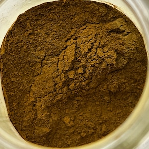May include: Close-up of a jar filled with a finely ground, dark brown powder. The powder has a textured appearance with visible cracks and variations in color, suggesting a natural product.