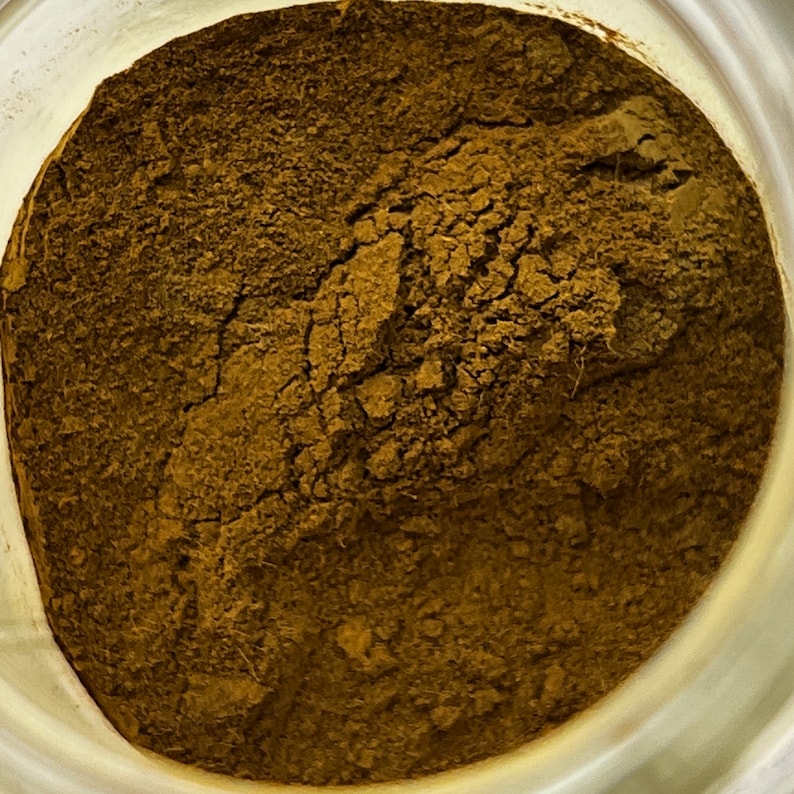 May include: Close-up of a jar filled with a finely ground, dark brown powder. The powder has a textured appearance with visible cracks and variations in color, suggesting a natural product.