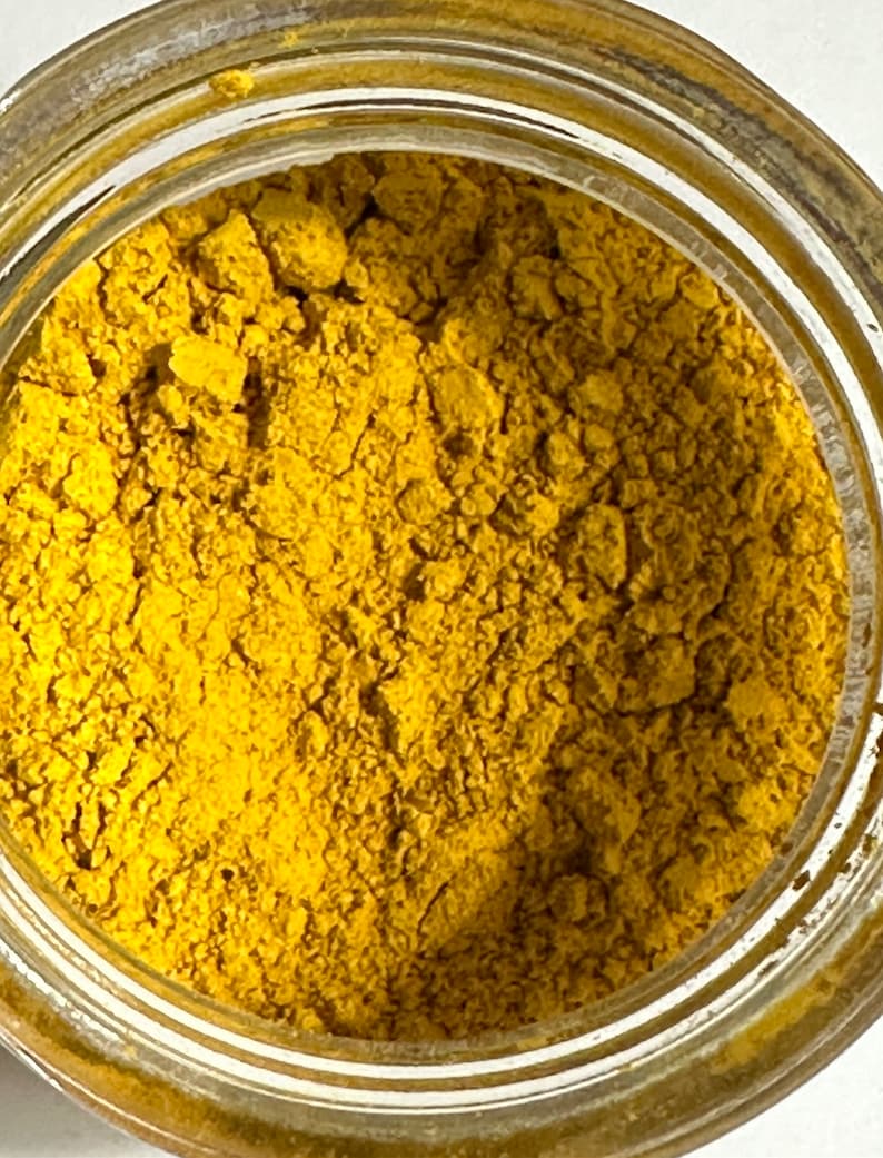 May include: Close-up of a clear glass jar filled with a vibrant yellow powder. The powder has a fine, granular texture and appears to be a spice or pigment. The jar's rim is visible, showing a metallic, aged finish. The overall image suggests a culinary or artistic product.