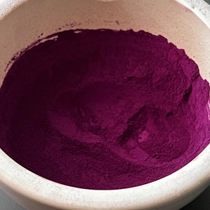 May include: A close-up shot of a white ceramic mortar filled with a vibrant, deep magenta powder. The powder fills the bowl, creating a textured surface. The mortar's rim is visible, contrasting with the rich color of the powder.
