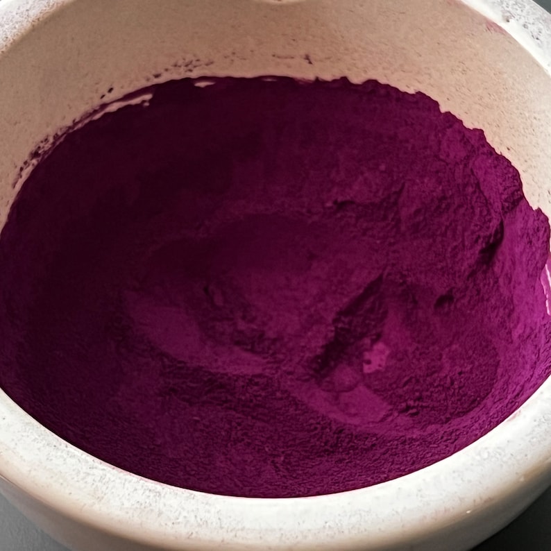 May include: A close-up shot of a white ceramic mortar filled with a vibrant, deep magenta powder. The powder fills the bowl, creating a textured surface. The mortar's rim is visible, contrasting with the rich color of the powder.