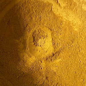 May include: Close-up of a pile of fine, golden-yellow powder. The powder has a textured surface with a slightly darker, central area. The overall appearance suggests a natural, earthy material, possibly a spice or pigment.