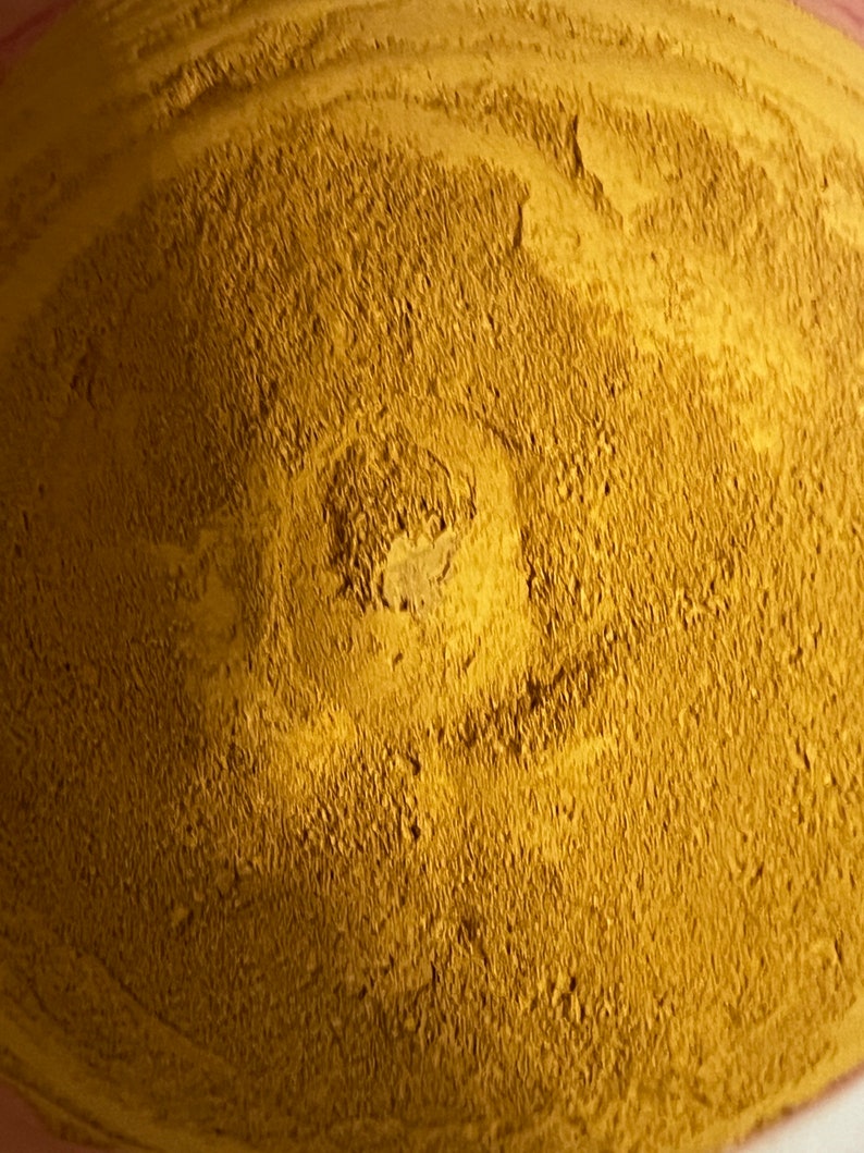 May include: Close-up of a pile of fine, golden-yellow powder. The powder has a textured surface with a slightly darker, central area. The overall appearance suggests a natural, earthy material, possibly a spice or pigment.