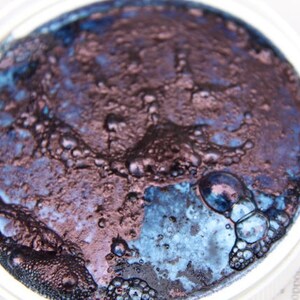 Puede incluir: Primer plano de un peque&ntilde;o recipiente de pintura met&aacute;lica azul oscuro y p&uacute;rpura con una superficie texturizada.