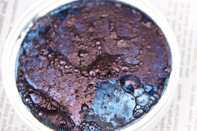 Puede incluir: Primer plano de un peque&ntilde;o recipiente de pintura met&aacute;lica azul oscuro y p&uacute;rpura con una superficie texturizada.