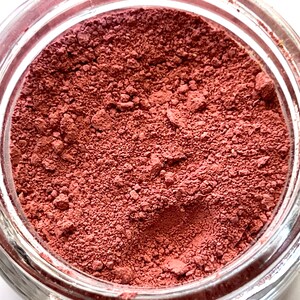 May include: A jar filled with a fine, reddish-brown powder. The powder has a slightly textured appearance, with small granules visible throughout. The glass jar is clear, allowing the color of the powder to be fully displayed. The contents appear to be a cosmetic product.