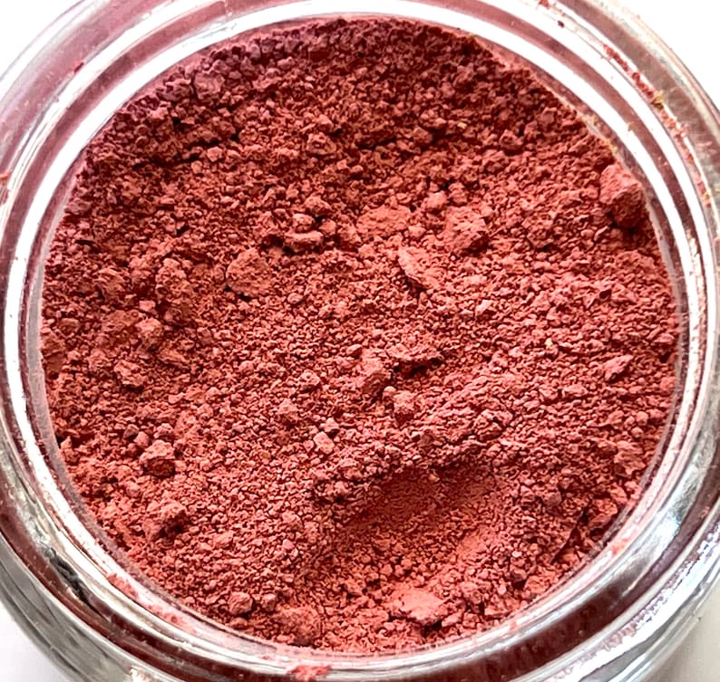 May include: A jar filled with a fine, reddish-brown powder. The powder has a slightly textured appearance, with small granules visible throughout. The glass jar is clear, allowing the color of the powder to be fully displayed. The contents appear to be a cosmetic product.