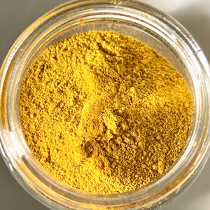 May include: A clear glass jar filled with a fine, golden-yellow powder. The powder has a textured appearance, filling the jar to the top. The jar's rim is visible, and the background is a soft, neutral tone.