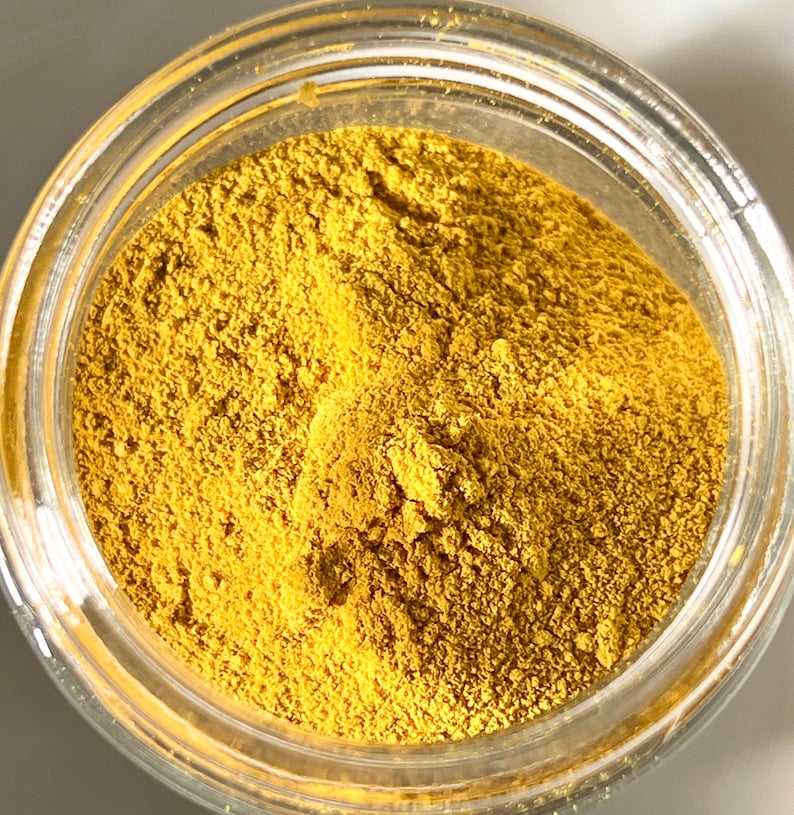 May include: A clear glass jar filled with a fine, golden-yellow powder. The powder has a textured appearance, filling the jar to the top. The jar's rim is visible, and the background is a soft, neutral tone.