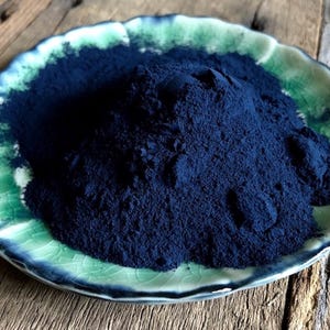 May include: A ceramic plate with a green and white rim holds a large pile of dark blue powder. The plate sits on a wooden surface. The powder appears finely ground and fills the center of the plate.