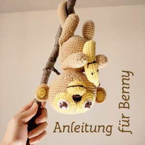 May include: A crocheted teddy bear toy in beige and yellow, hanging upside down from a tree branch. The bear has brown and white eyes and is holding a yellow banana. The text "Anleitung für Benny" is visible.