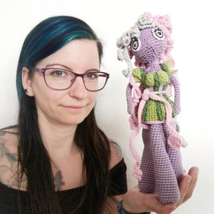 Peut inclure: Poupée au crochet faite à la main avec un corps lavande, des accents verts et roses et des cheveux gris. La poupée porte un haut vert et des rubans roses. La poupée est tenue dans la main d'une personne.