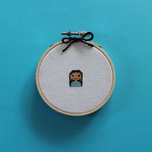 Puede incluir: Un pequeño aro de bordado redondo con un fondo de tela blanca. La tela tiene un diseño de punto de cruz de una persona con cabello castaño y una camisa azul.