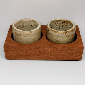 May include: Two small, cylindrical ceramic cups with speckled, off-white exteriors sit in a rectangular, reddish-brown wooden tray. The cups have a rustic, handmade appearance, perfect for serving small portions.