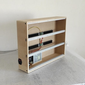 Puede incluir: Una caja de madera con dos estantes y una fuente de alimentación en su interior. La caja tiene un cable de alimentación negro y un cable rojo y negro que va a los estantes. Los estantes son de metal y tienen un acabado plateado.