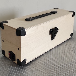 Può includere: Una scatola rettangolare in legno chiaro dotata di maniglia superiore nera, angolari di protezione in metallo nero e una chiusura frontale nera. Il lato della scatola presenta una porta di alimentazione nera con un piccolo interruttore rosso.