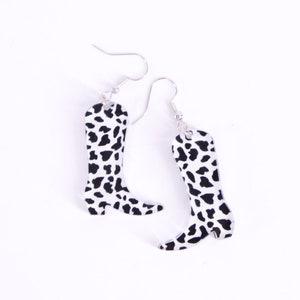 May include: A pair of silver dangle earrings featuring a cow print design in the shape of a cowboy boot.
