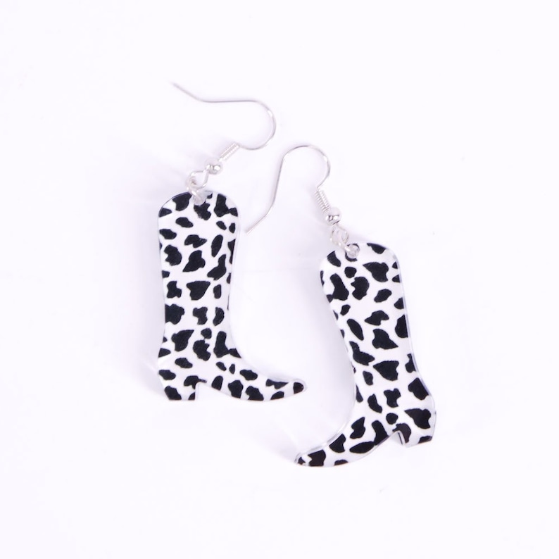 May include: A pair of silver dangle earrings featuring a cow print design in the shape of a cowboy boot.