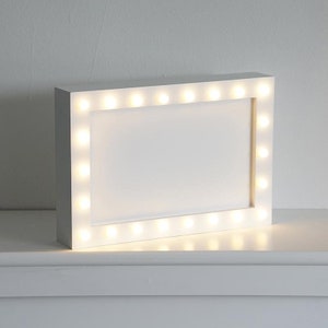 May include: White rectangular picture frame with a white border and a row of small, warm white LED lights around the edge.