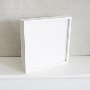 May include: A square shadow box frame with a clean white finish and a deep profile. The frame features a smooth, matte white border surrounding a blank, flat white interior panel, presented in a simple, unadorned, and functional design.
