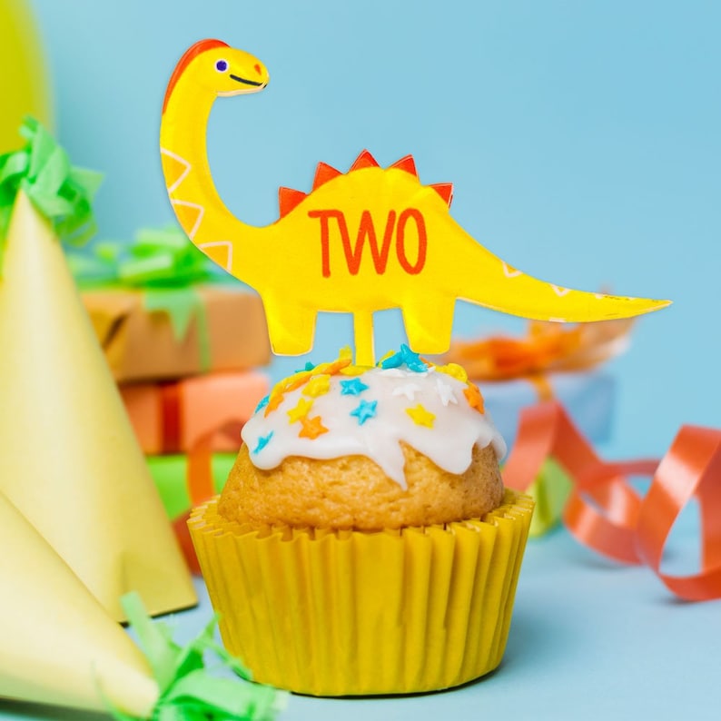 May include: A yellow cupcake with white frosting and colorful sprinkles topped with a yellow dinosaur cake topper that says "TWO" in red letters.