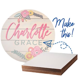May include: A white wooden round sign with a gray and white woodgrain pattern. The sign has a pink floral wreath with a pink and white floral design. The text "Charlotte Grace" is written in pink and gray letters on the sign.