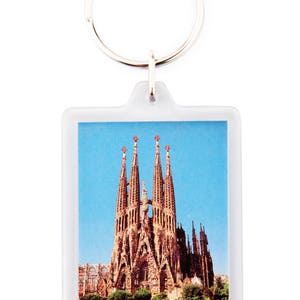 May include: A white plastic keychain with a picture of the Sagrada Familia church in Barcelona, Spain. The church is a famous landmark with tall spires and intricate details.