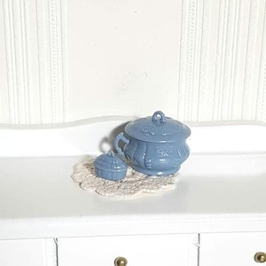May include: A light blue ceramic sugar bowl with a lid and a small matching creamer, both with crown details. They sit on a white crocheted doily. The set is displayed on a white surface.