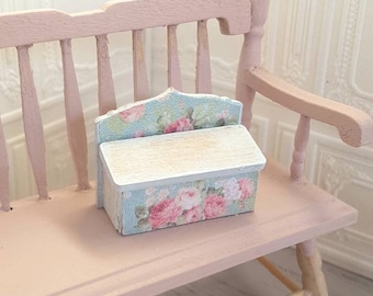 May include: A miniature pink bench with a small box featuring a floral pattern. The box has a light blue background with pink roses and green leaves. The bench has a slatted back and armrests, and is painted in a soft pink color.