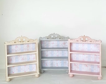 May include: Three miniature wooden shelves with ornate tops. The shelves are painted in white, gray, and pink. Each shelf has three tiers and is lined with a floral patterned paper.
