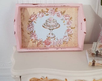 May include: A pink wooden tray with a distressed finish. The tray features a printed design of a cake with roses around it. The tray is sitting on a white dresser with gold accents.