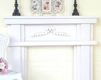 May include: White painted wooden fireplace mantel with a decorative leaf design. The mantel has a shelf and is set against a white wall with decorative molding.