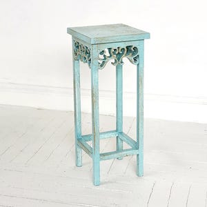 May include: A light blue wooden plant stand with intricate carved details and a square top. The stand has a distressed finish and is supported by four legs.