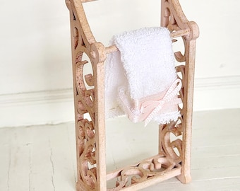 Può includere: Un portasciugamani in legno rosa con tagli decorativi. Un asciugamano bianco è appeso alla barra.