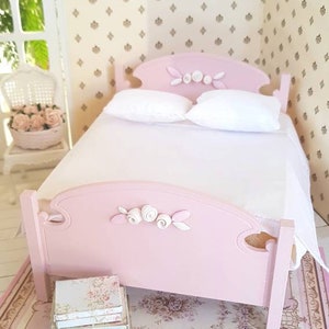 May include: A pink dollhouse bed with white bedding and a floral patterned rug. The bed has a white rose detail on the headboard.