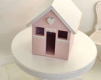May include: A small pink wooden house with a white roof and a heart cutout on the front. The house has white lace trim around the windows and a white heart on the roof.