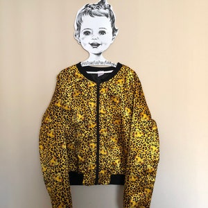 May include: A yellow and black bomber jacket with a leopard print and cartoon lion faces. The jacket has a zipper closure and black ribbed cuffs and waistband.