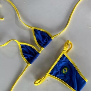 Puede incluir: Un conjunto de bikini azul y amarillo. La parte superior e inferior del bikini son azules con ribetes y correas amarillas. La parte inferior presenta un pequeño emblema de la bandera brasileña.