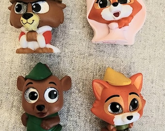 Puede incluir: Cuatro pequeñas figuras de plástico de personajes de la película de Disney Robin Hood. Los personajes son Robin Hood, Little John, Maid Marian y el Príncipe Juan. Las figuras están todas en diferentes poses y tienen ojos grandes y expresivos.