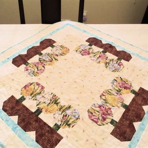 May include: A square table runner with a light beige background and a blue border. The runner features a repeating pattern of brown flower pots with colorful flowers.