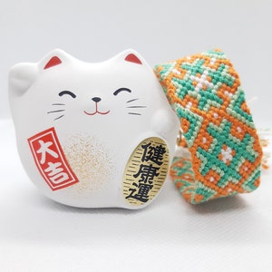May include: White ceramic cat figurine with a raised right paw, a red and gold label that says "大吉" and a gold label that says "招福", and a green and orange woven friendship bracelet.