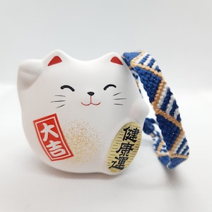 Könnte beinhalten: Weiße Keramik-Katzenfigur mit einem blau-weißen geflochtenen Band. Die Katze hat einen roten Mund und rote Ohren. Die Figur ist mit goldenem Glitzer verziert und trägt den Text "大吉" und "招福" in Schwarz.