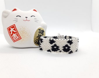 Puede incluir: Una pulsera de amistad tejida en blanco y negro con un patrón geométrico se muestra junto a una figura de gato Maneki-neko de cerámica blanca. El gato tiene un collar rojo y una moneda de oro.