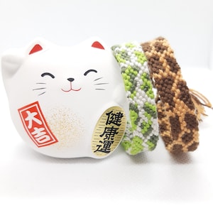 May include: A white ceramic Maneki-neko cat figurine with red accents and Japanese text, next to two woven friendship bracelets. One bracelet is green and white, the other is brown with a giraffe pattern.